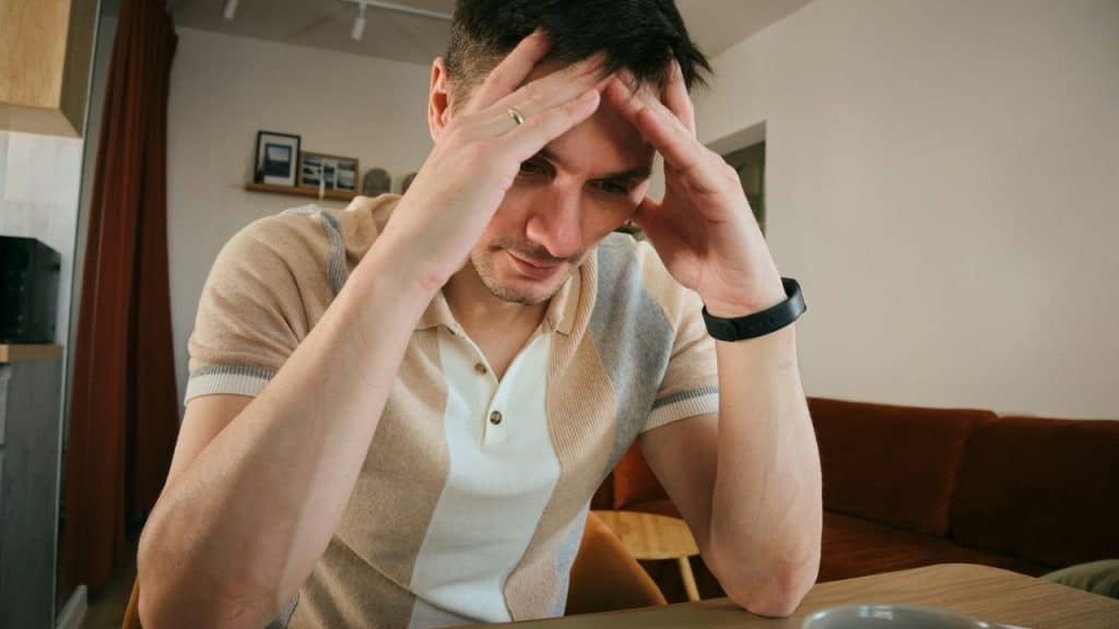 A man in a tan polo shirt sits at a table holding his forehead.