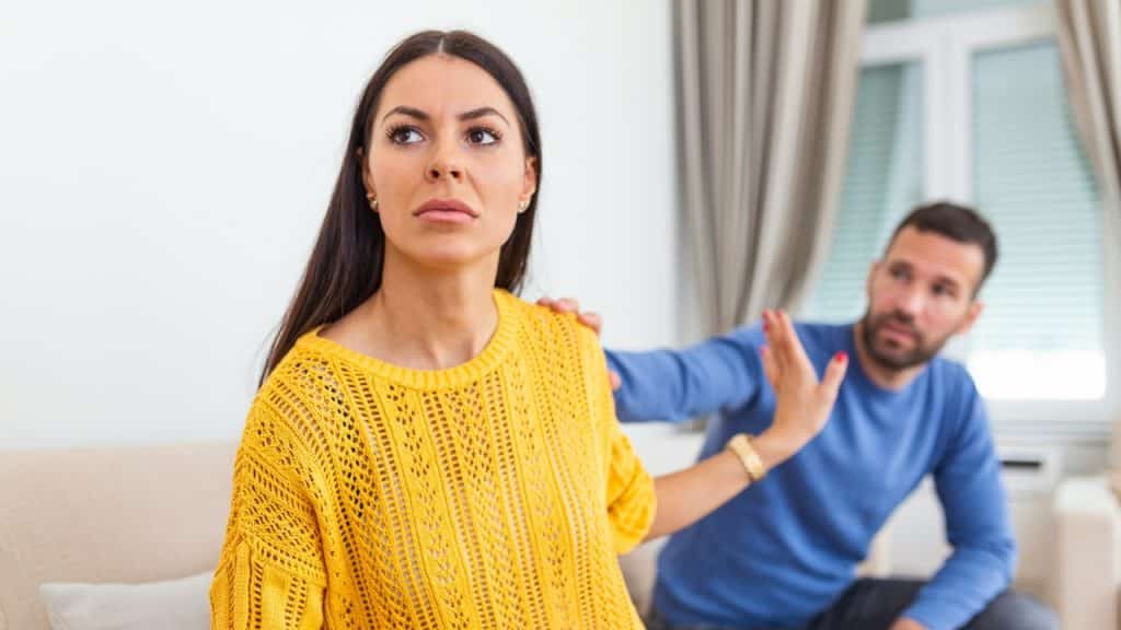 A woman in a yellow sweater holds up a hand to a man sitting behind her.