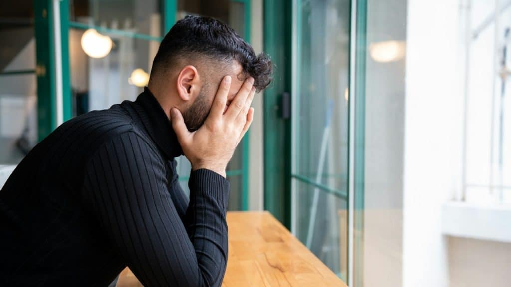 A man in a black turtleneck sits at a table, covering his face with both hands.