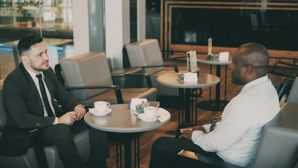 Two men in business attire sit at a table in a cafe having a conversation.