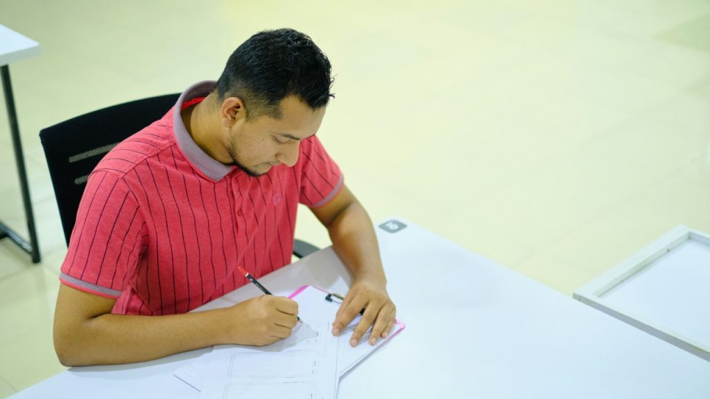 A man in a red polo shirt writes on a paper placed on a white desk.