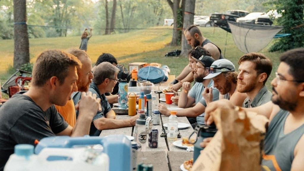 A group of men sit at a long wooden picnic table eating outdoors during daytime.