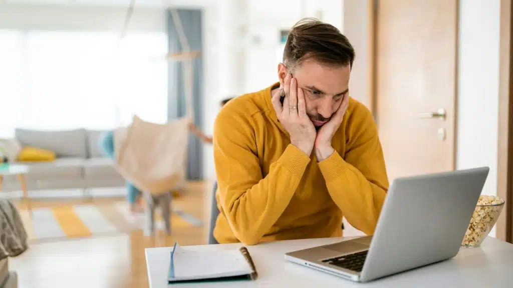A frustrated man in a yellow sweater looks at a laptop, holding his face in his hands.