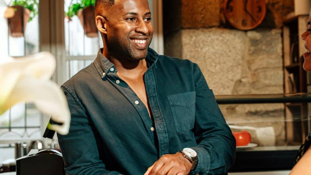 A smiling person in a dark shirt chatting at a cozy kitchen counter.