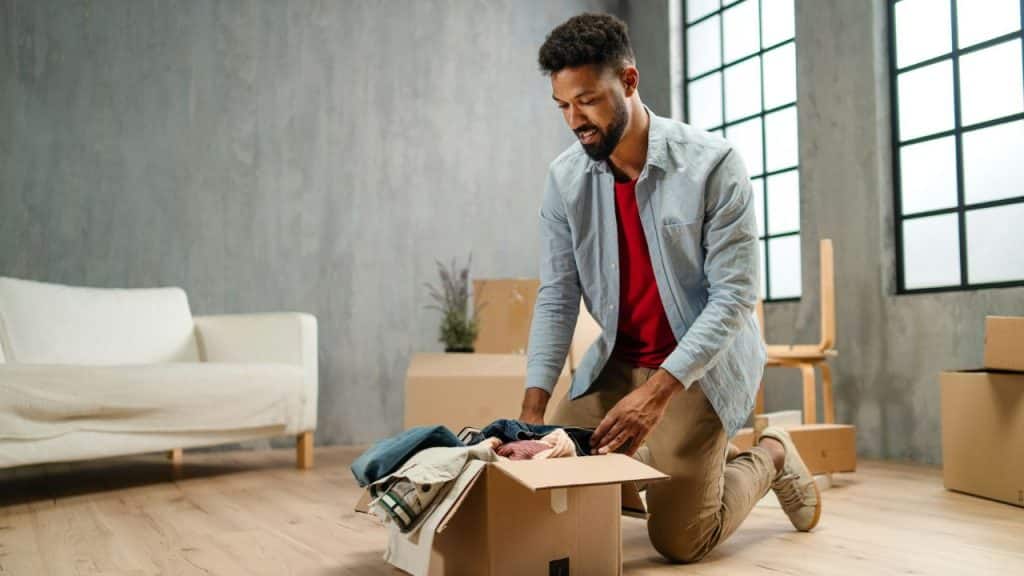 A man kneels on a wooden floor, packing or unpacking clothes into a cardboard box.