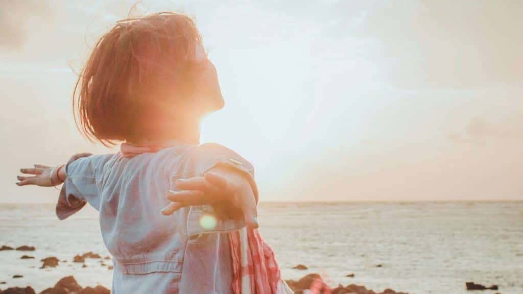 A woman standing by the sea with her hands outstretched as the sun nears setting in the sky.