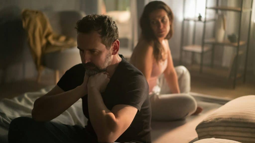 A woman looking at a man who is sitting with his back to her and holding his neck with his hands.