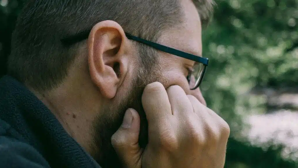 A man wearing glasses touching his beard outdoors.
