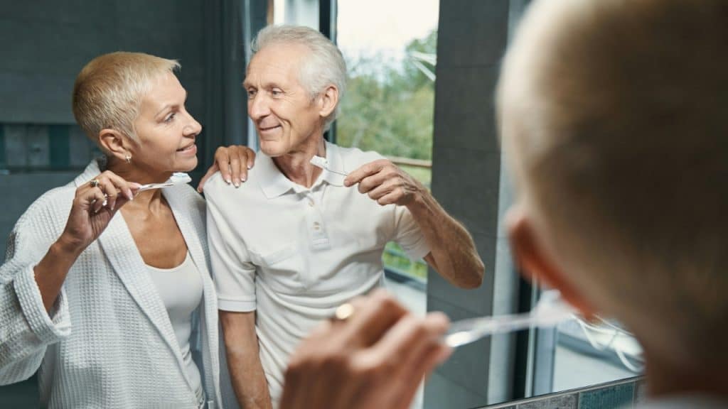 An older man and woman brush their teeth together while looking into a bathroom mirror.