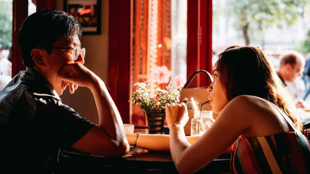 A man and woman sit at a table facing each other in warm, natural light.