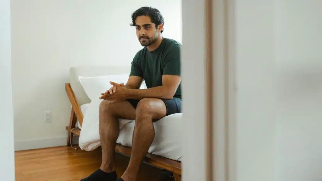 A man in a dark t-shirt sits on the edge of a bed with his hands clasped.