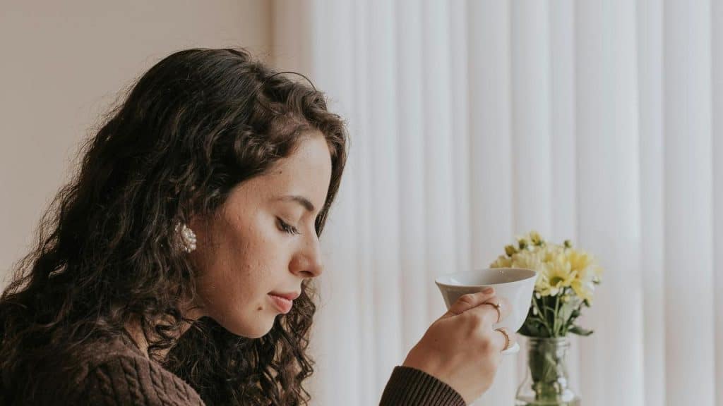 A woman holding a cup and looking down beside a vase of flowers indoors.