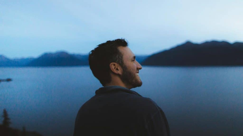 A man smiling in profile while looking out over a calm lake at dusk.