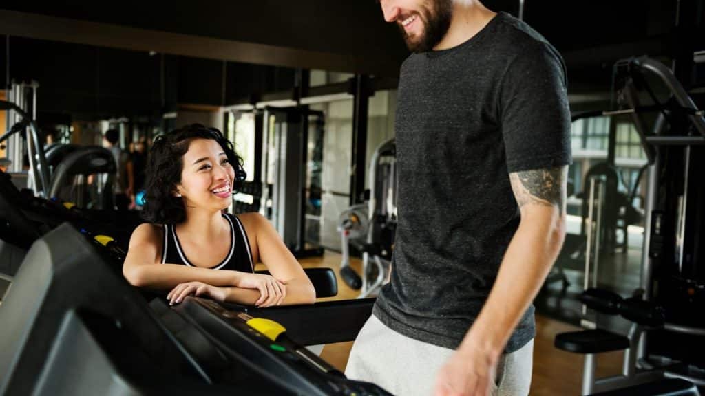 A woman smiling at a man while resting on a treadmill in a gym.