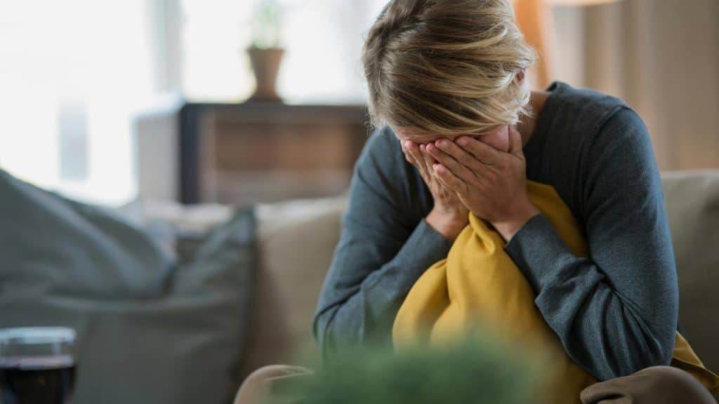 A person sitting on a couch, covering their face with their hands while holding a pillow.