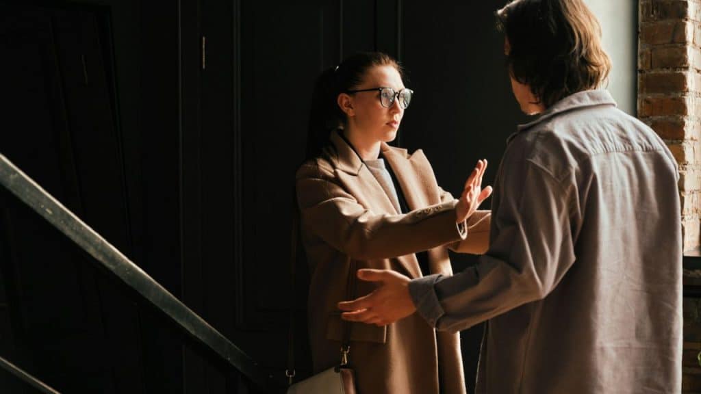 A woman in a tan coat and glasses holds her hand up, facing a person with their back to the camera, in an indoor setting.