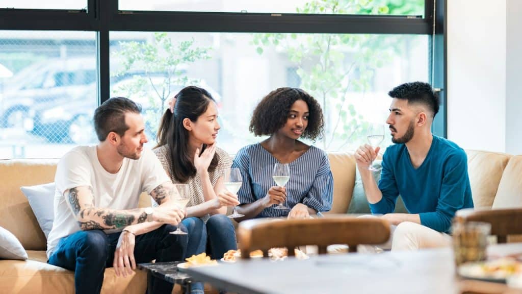 Four people sit on a couch together indoors holding wine glasses while engaged in conversation.