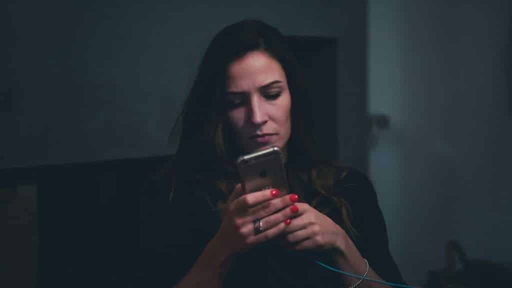 A woman with red nail polish looks intently at her phone in a dimly lit room.