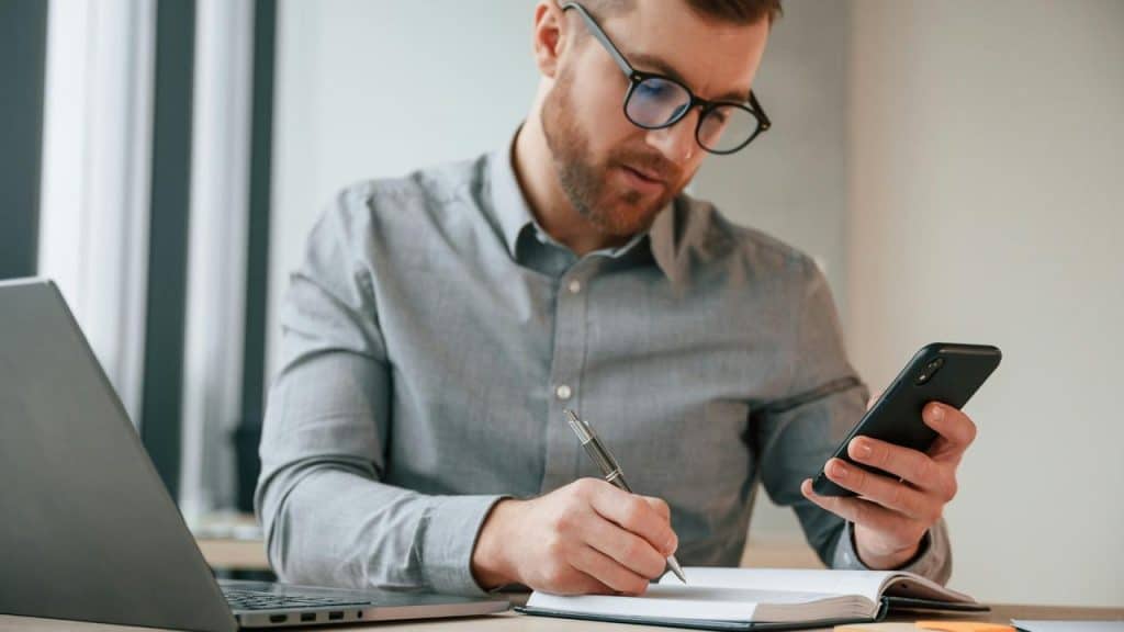 A man wearing glasses writing in a notebook while checking his phone at a desk.