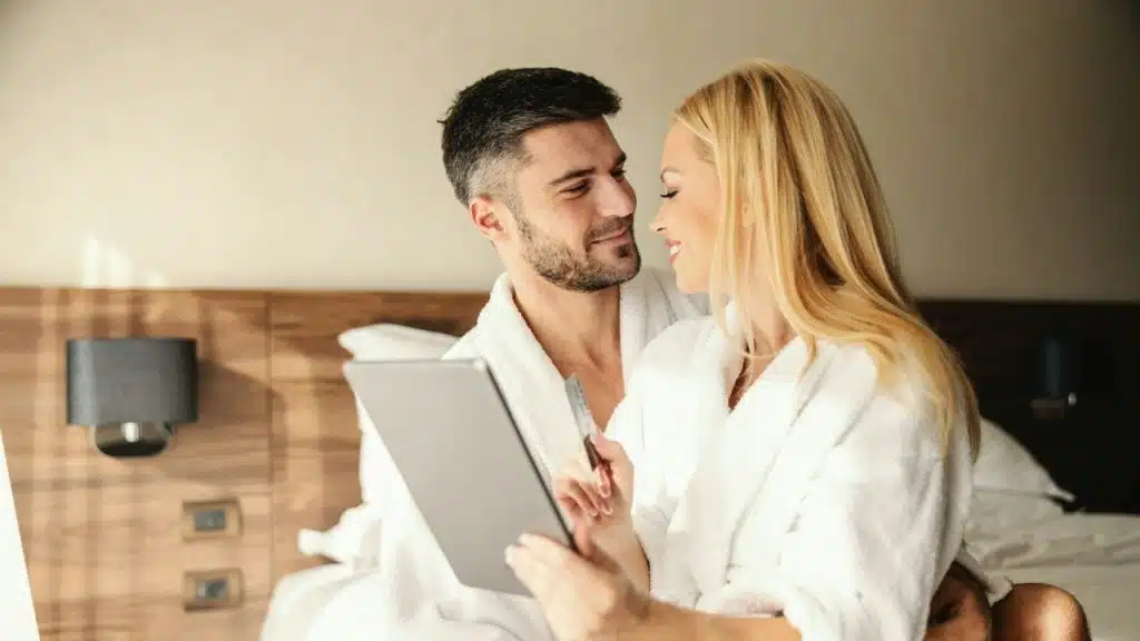 A man and woman in white robes sitting on a bed looking at a digital tablet.