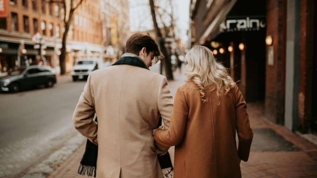 A man and woman in tan coats walk down a city sidewalk away from the camera.