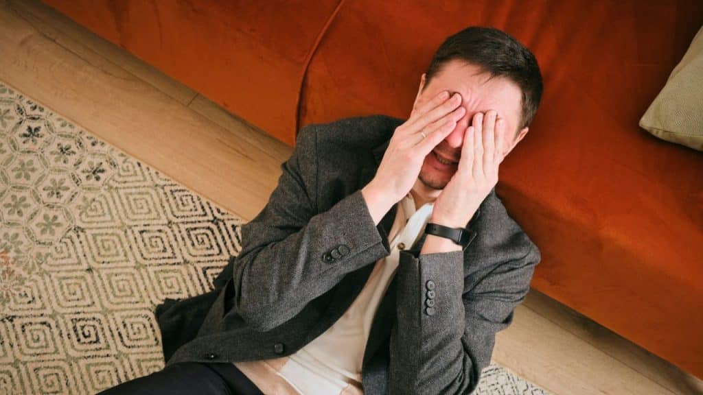 A man in a blazer sits on the floor covering his eyes with both hands.
