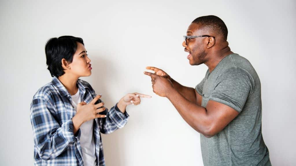 A man and woman gesture and point at each other against a plain white wall.