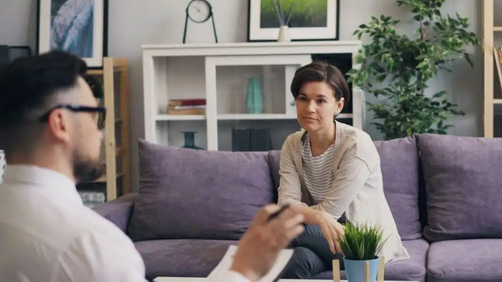 A woman sits on a purple sofa and listens to a man holding a pen.