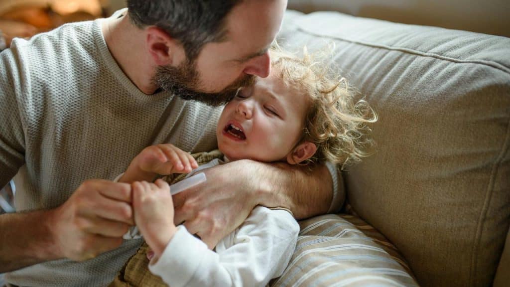 A father comforting a crying toddler while giving medicine.