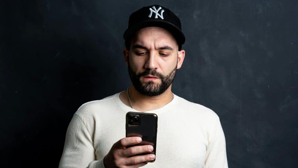 A man in a baseball cap looks down at his phone against a dark background.