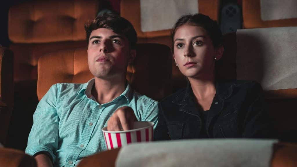 A young couple sitting in a movie theater, watching a film with a bucket of popcorn.