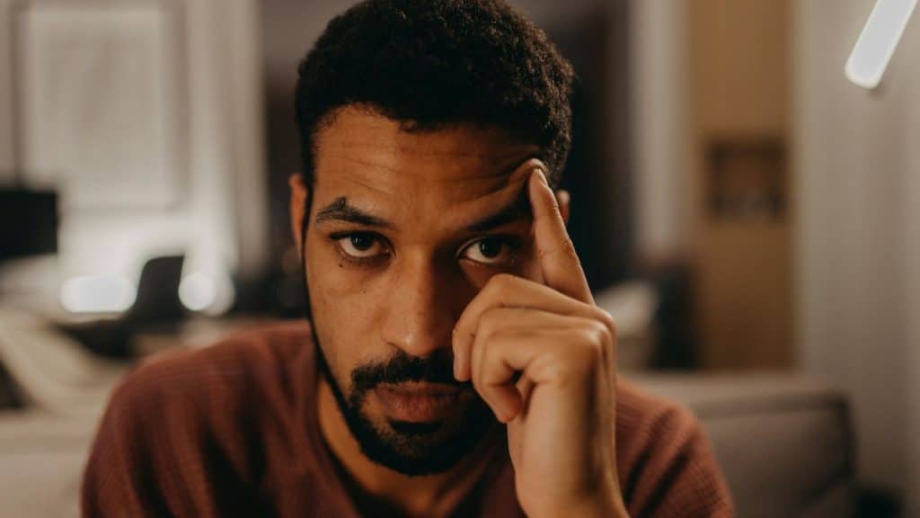 A man sits in low light with his finger pressed against his temple looking forward.