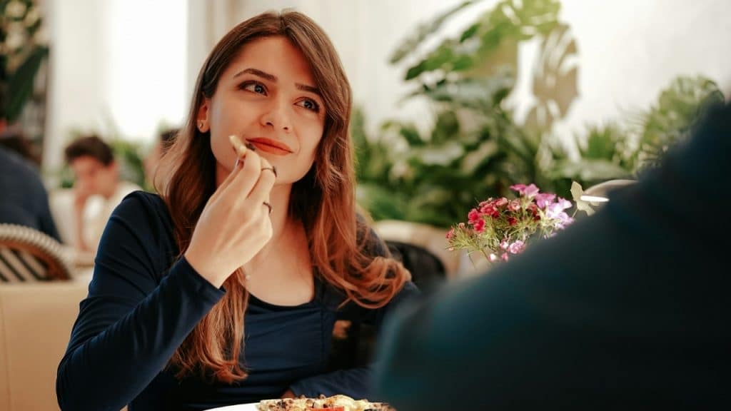 A woman in a blue dress eats a piece of food while looking at someone off-camera.