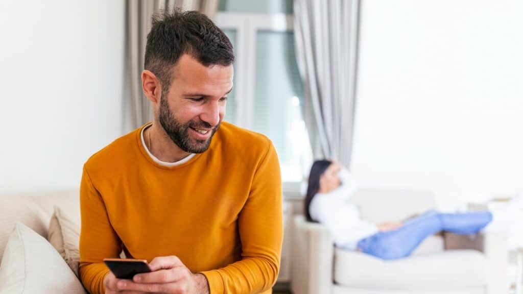 A man smiling at his phone sits on a couch while a woman sits in background.