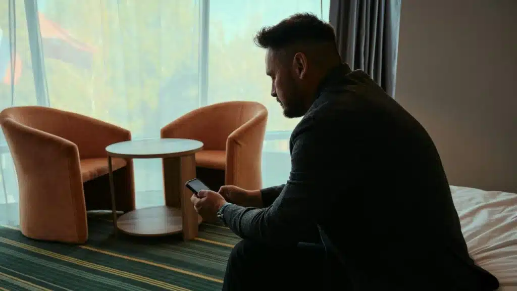 A man sits in profile on a bed, looking down at a smartphone in his hands.