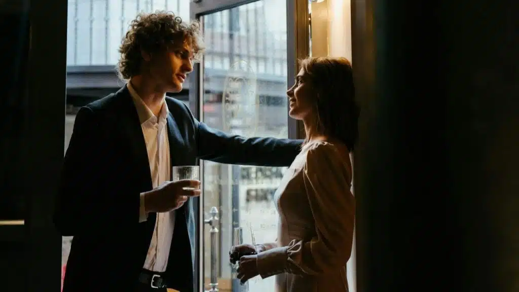 A man and woman holding drinks stand near a glass door and look at each other.
