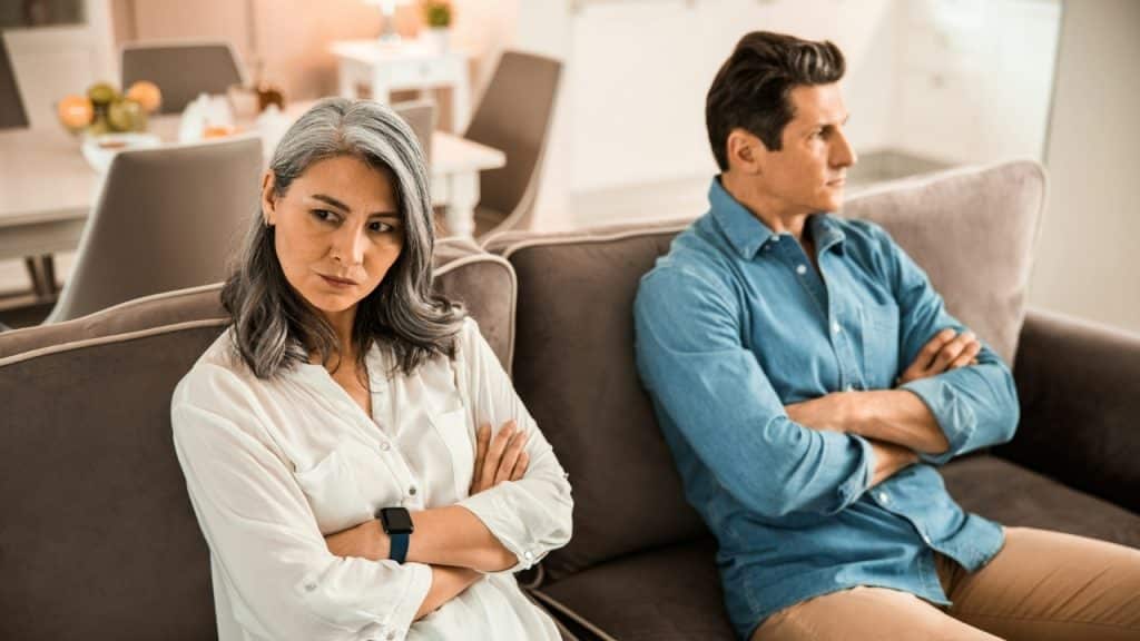 A man and woman sit on a sofa with crossed arms, looking away from each other.