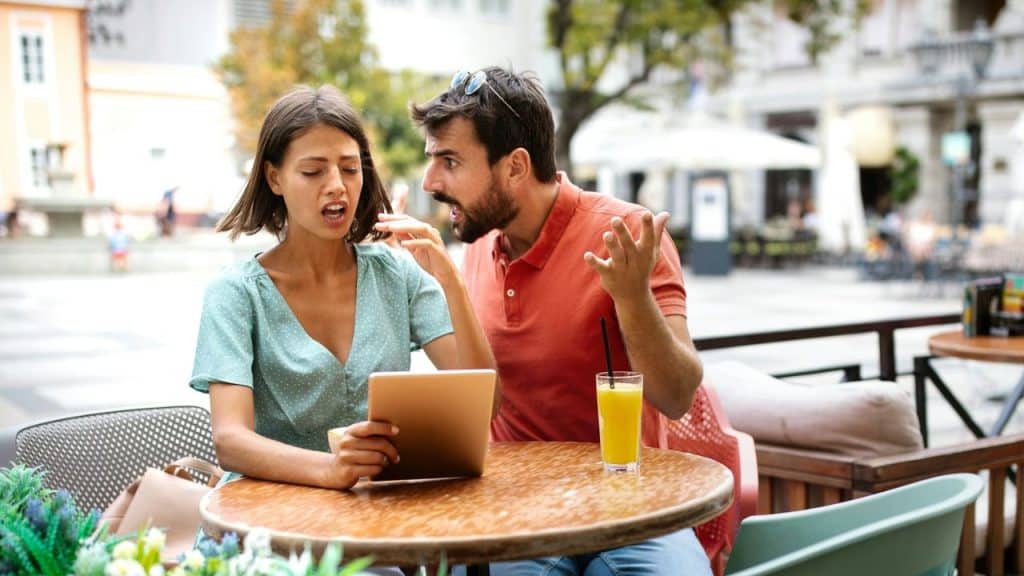 A woman holds a tablet while a man beside her gestures and speaks with intensity.