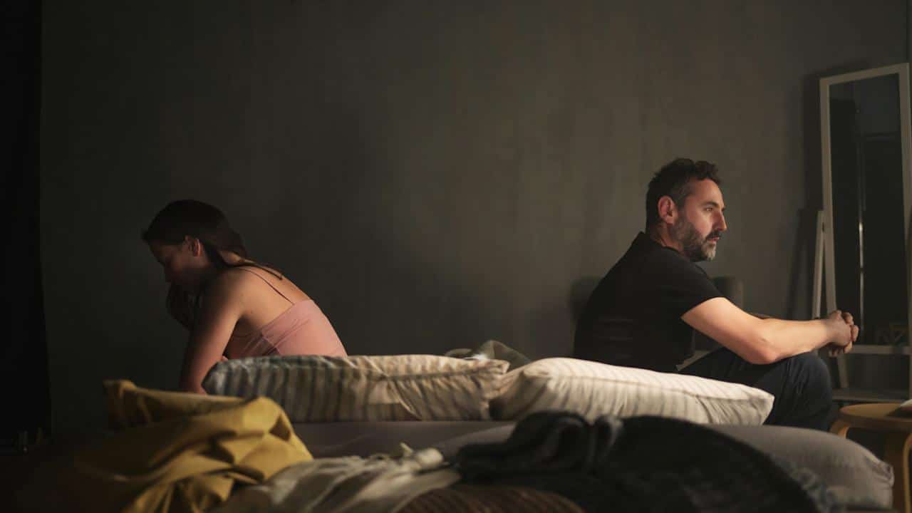 A man and a woman sit facing away from each other on a bed in a dimly lit room.