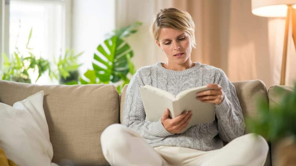 A woman with short blonde hair sits on a beige sofa and reads a book.