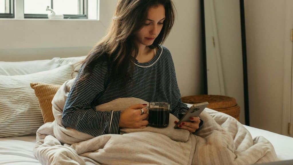 A woman in a striped shirt sitting in bed with coffee and her phone on a sunny morning.