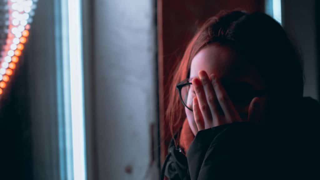 A person covering their face with both hands in a dimly lit indoor setting.