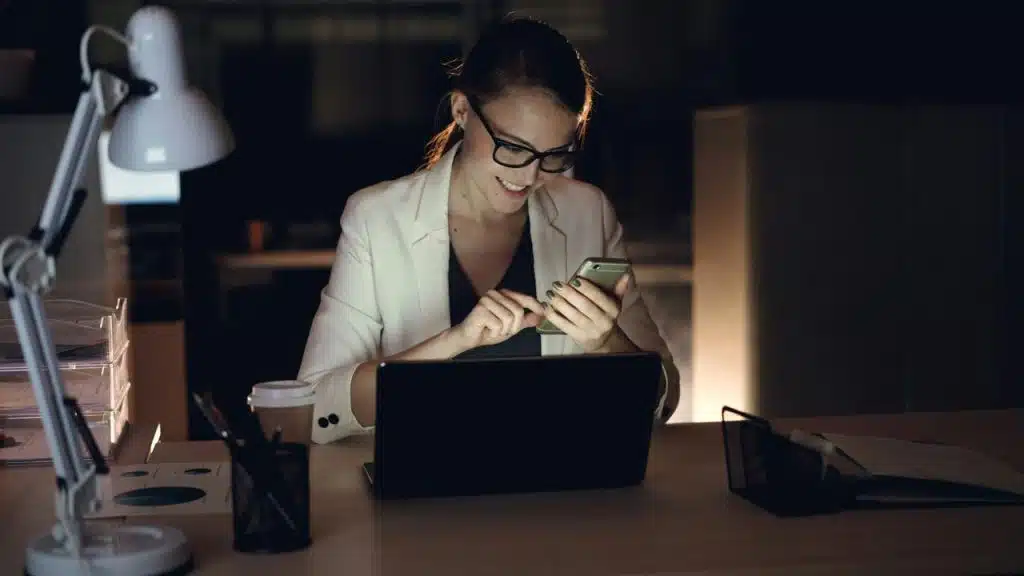 A woman smiling while using a smartphone and laptop at a desk.