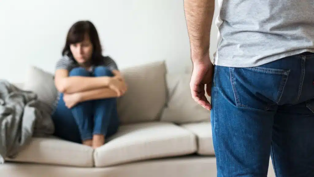 A man stands in the foreground with his back turned toward a woman sitting on a sofa.
