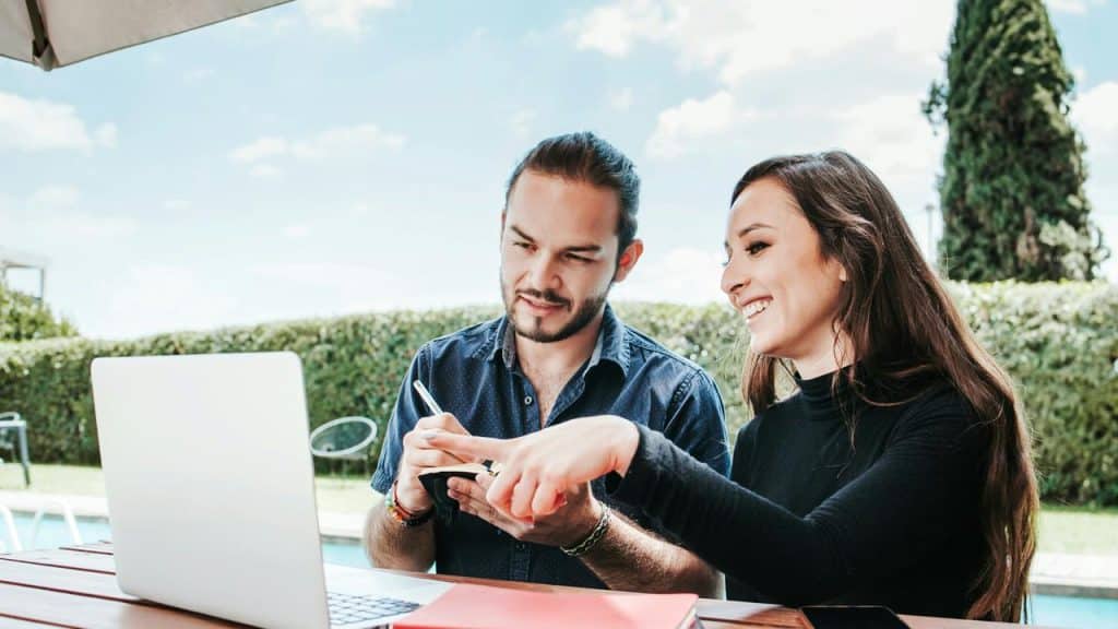 A couple working together on a laptop at an outdoor table on a sunny day.