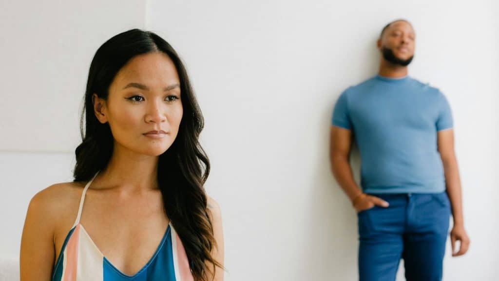 A woman looks forward somberly while a man stands blurred in the background against a white wall.