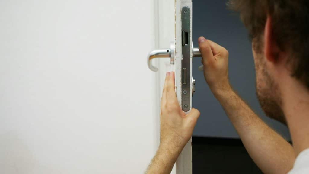 A close-up shot shows a man's hands installing or repairing a metal door handle mechanism.