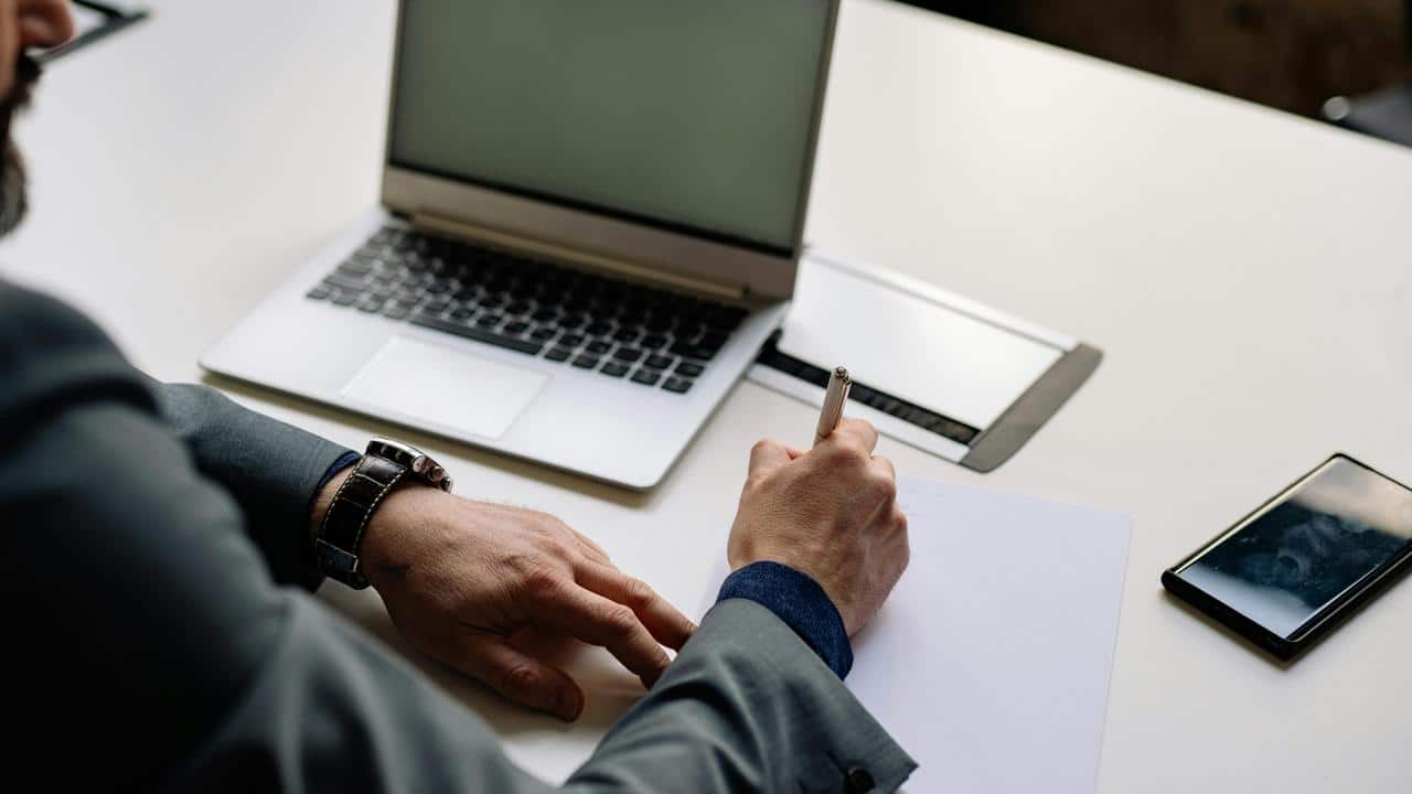 A person writing on paper next to a laptop and phone.