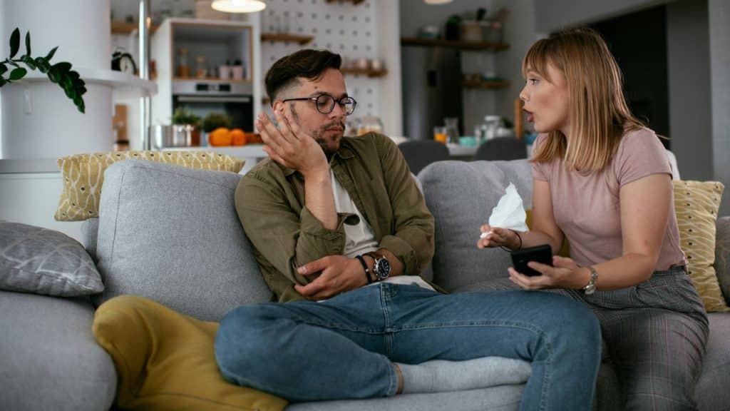 A woman speaks and holds a tissue while a man sits beside her looking away.