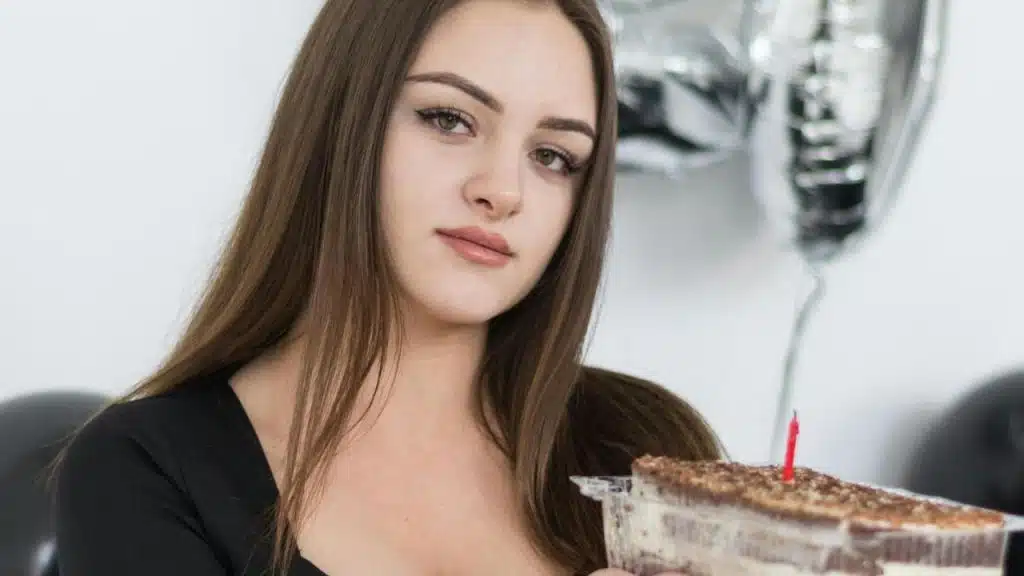 A woman with long brown hair holds a cake with a single red candle while looking forward.